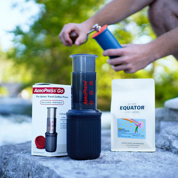 An Aeropress Go with a bag of coffee and a person using a manual grinder in the background.