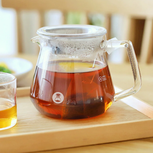 Clear glass server with tea steeping in it, on a wooden board.
