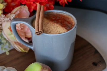 Blue mug with apple cider. A cinnamon stick on the mug and blurred flowers in the background. A part of an apple in front of the mug.