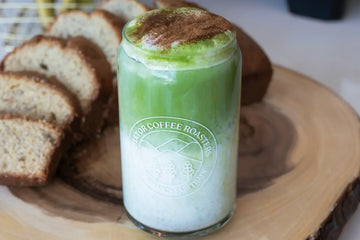Banana Bread iced matcha in a glass with banana bread behind it on a wooden surface.