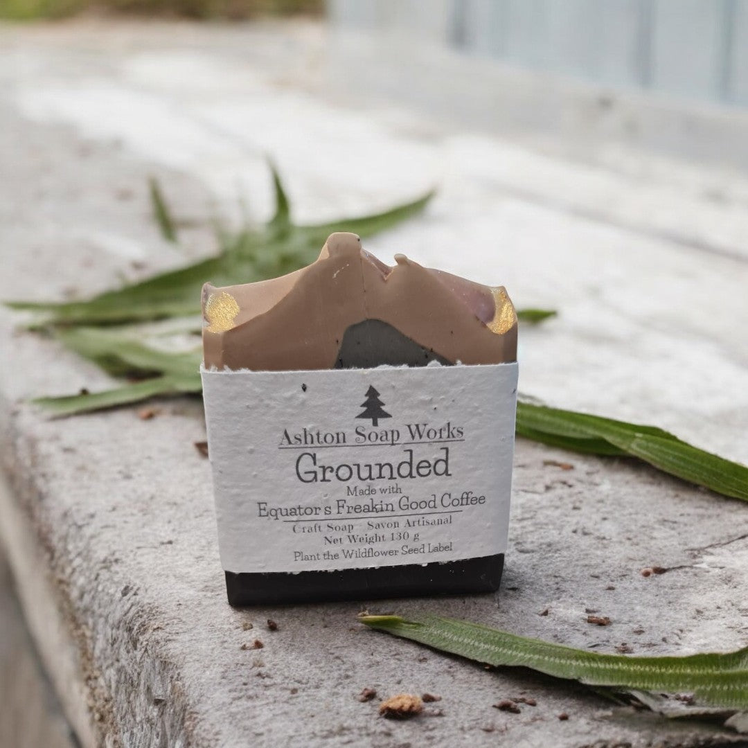 A bar of Ashton Soap Works Grounded coffee infused soap placed on a surface with leaves around it, showing the product label.