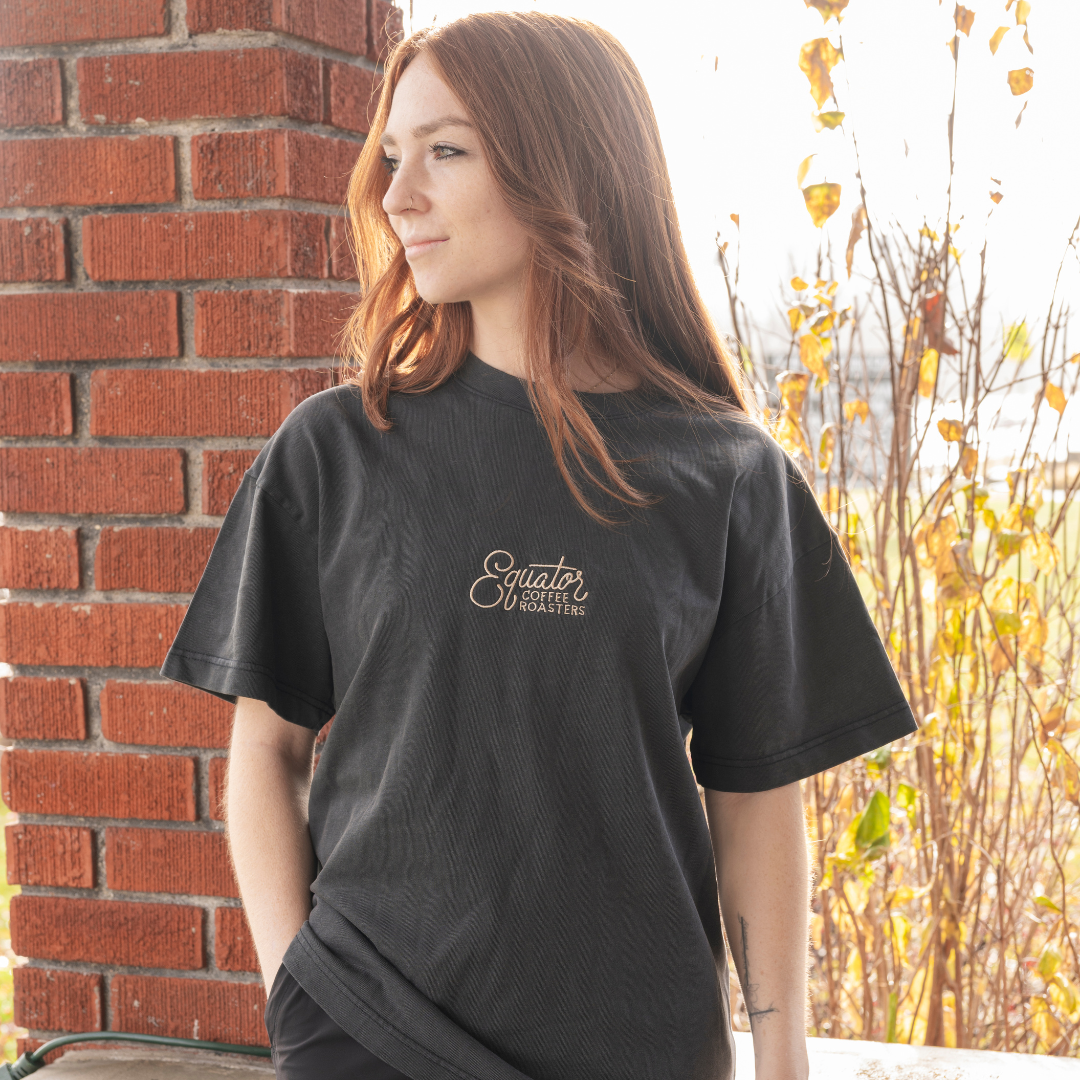 Person wearing a black t-shirt with a logo, standing against a brick wall with autumn foliage.