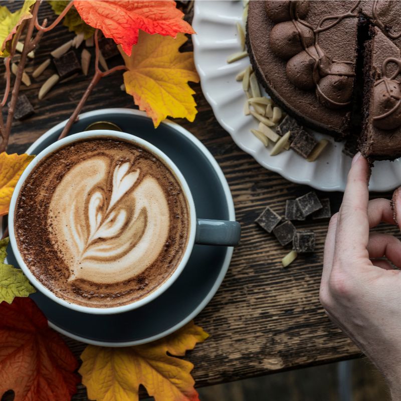 Chocolate cake with a slice cut, next to a cup of coffee with latte art on a wooden table with autumn leaves.