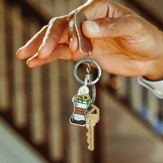 Hand holding a keychain with a key and Blooming Chemex charm against a blurred background
