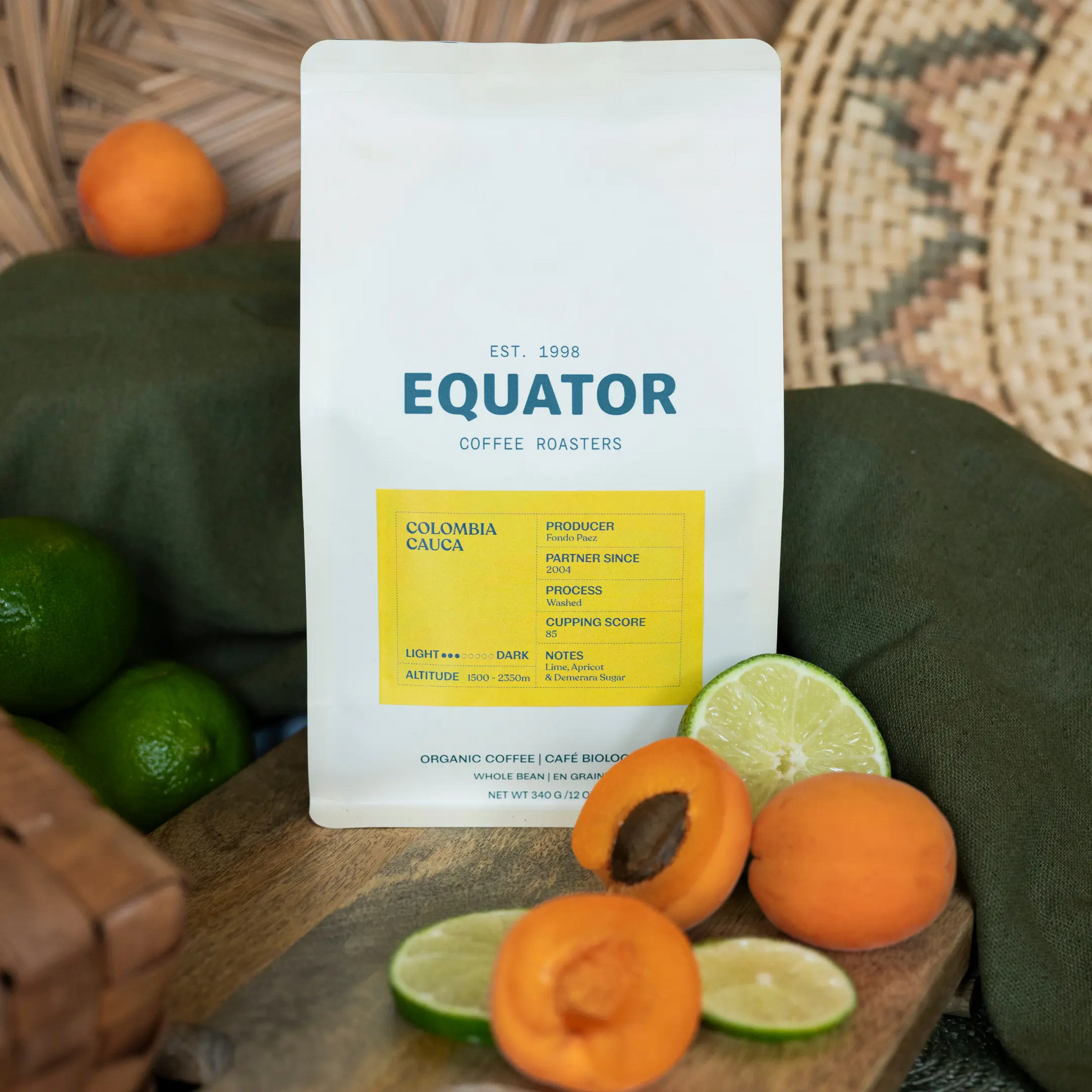 A 340g package of Colombia Cauca coffee beans stands on a wooden board with sliced limes, apricots, and whole fruit around it. The package shows the brand name EQUATOR COFFEE ROASTERS, plus a yellow label panel.