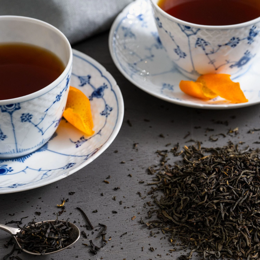 Two cups of tea with saucers, dried tea leaves, and orange slices on a dark table.