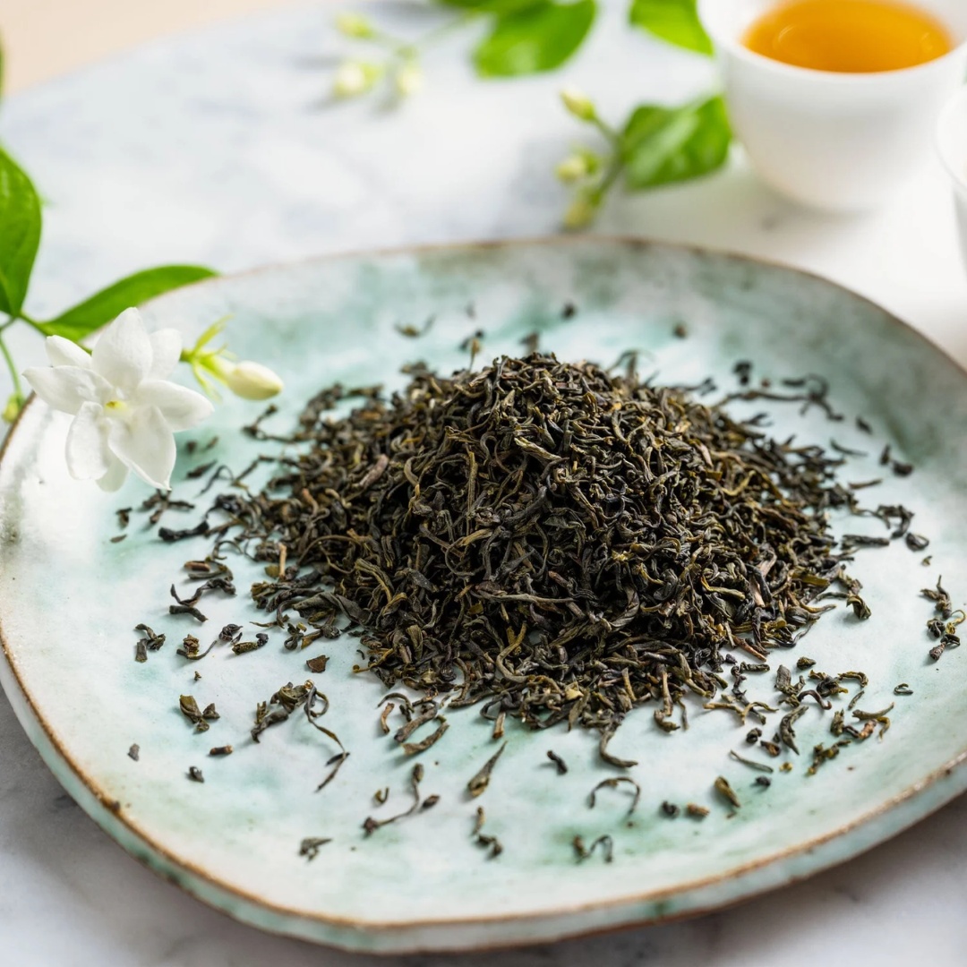Jasmine green tea leaves on a ceramic plate with a small cup of tea in the background.