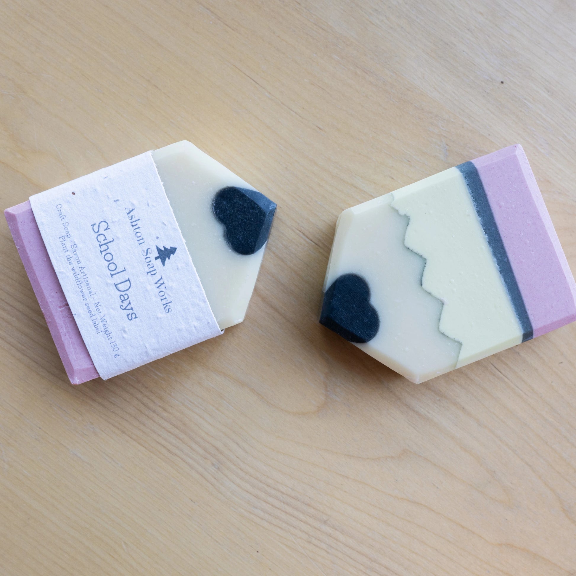 Two handmade soap bars shaped like pencils on a light wood background. One bar is wrapped in a white paper label that reads 'Ashton Soap Works School Days,' and the other is unwrapped to show a cream body with a black tip, a pink bottom, and a dark blue vertical stripe.