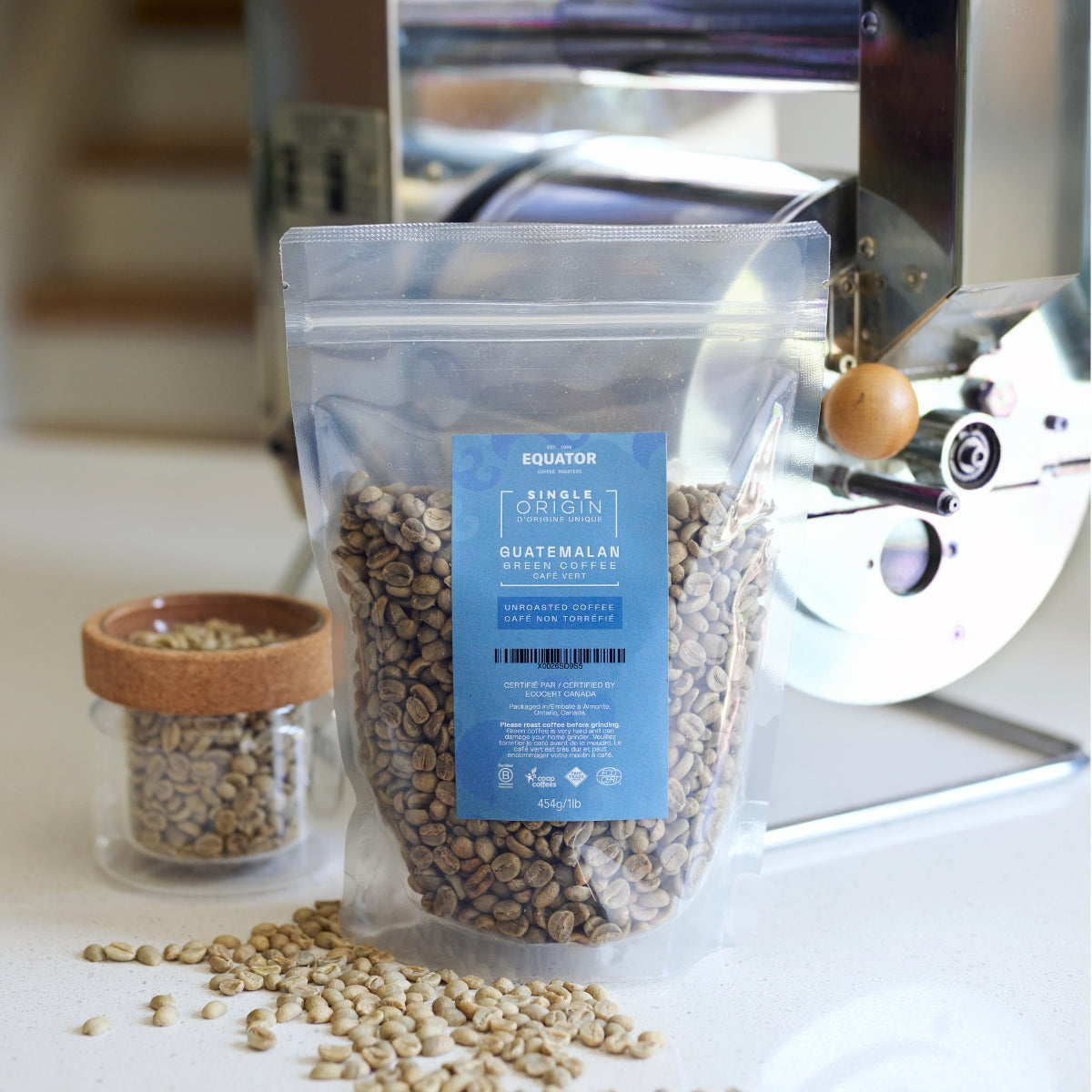 1lb bag of green unroasted coffee beans. There are loose coffee beans scattered on the surface next to the bag, and a small container and small home roaster in the background
