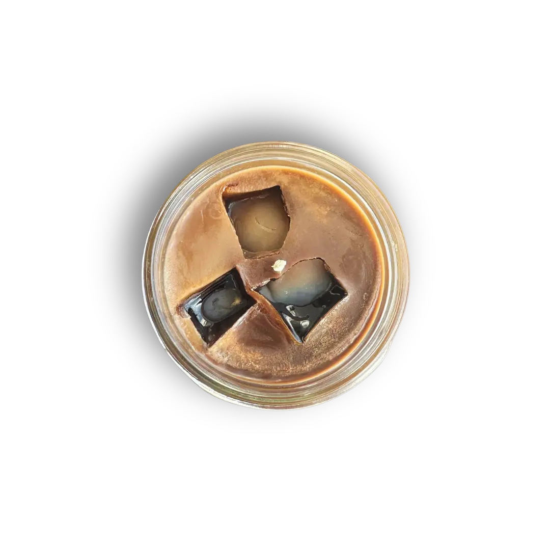 A top-down view of a clear glass jar containing a brown candle with three broken, glossy black square wax pieces embedded in the surface to look like ice cubes.